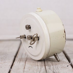 May include: A white vintage alarm clock with a silver winding key and a silver knob on the side. The clock is lying on a wooden surface.