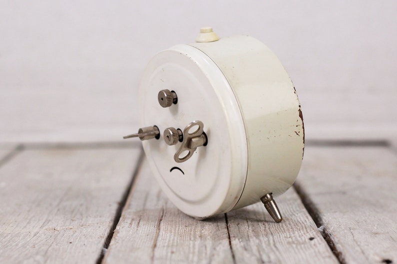 May include: A white vintage alarm clock with a silver winding key and a silver knob on the side. The clock is lying on a wooden surface.