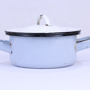 May include: A small, light blue enamel pot with a white lid and two side handles. The pot has a black rim and a white handle on the lid. The pot is likely vintage and shows signs of wear.