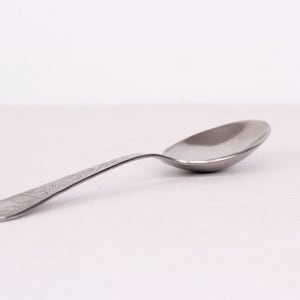 May include: A silver spoon with an ornate design on the handle. The spoon is lying on a white surface.