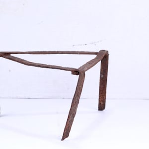 Può includere: Un cavalletto in metallo arrugginito, probabilmente utilizzato per sostenere una pentola o un bollitore sopra un fuoco.