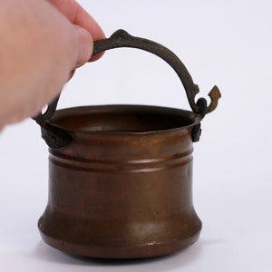 May include: A small copper pot with a handle. The pot is brown and has a shiny finish. The handle is made of metal and is attached to the pot with rivets.