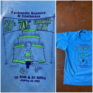 May include: A light blue t-shirt with a cartoon illustration of a person running around a birthday cake. The text on the shirt reads "Panhandle Runners & Triathletes ED's 70th Birthday 7K Run & 5K Walk January 30, 1993".