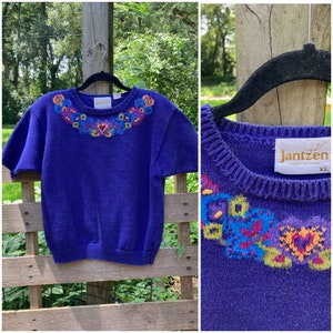 Puede incluir: Su&eacute;ter morado de manga corta con un cuello bordado con flores de colores. El su&eacute;ter tiene una etiqueta Jantzen con el texto "Jantzen Made in China XL".