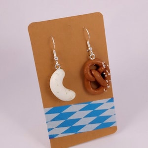 Könnte beinhalten: Ein Paar Hängeohrringe mit einem weißen Brezel und einem braunen Brezel mit weißen Streuseln. Die Ohrringe werden auf einer braunen Karte mit einem blau-weiß karierten Muster präsentiert.