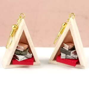 Könnte beinhalten: Ein Paar dreieckige Ohrringe mit einem Miniatur-Bücherregal-Design. Jeder Ohrring zeigt einen Stapel von fünf Miniaturbüchern mit einem roten Buch unten.