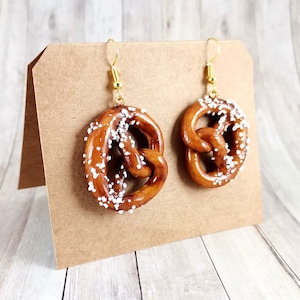 Op de afbeelding: Een paar gouden oorhangers met een hanger in de vorm van een pretzel met witte sprinkles. De oorbellen worden gepresenteerd op een bruine kaart.