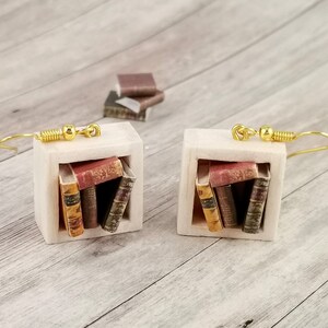Könnte beinhalten: Ein Paar goldene Ohrringe mit einem Miniatur-Bücherregal-Design. Jedes Bücherregal verfügt über einen Stapel von fünf Miniaturbüchern mit roten, braunen und goldenen Einbänden.