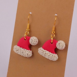 Könnte beinhalten: Ein Paar festliche Ohrringe mit Miniatur-Weihnachtsmützen. Die Mützen sind leuchtend rot mit weißem, flauschigem Besatz und einem kleinen weißen Bommel. Die Ohrringe hängen an goldfarbenen Fischhaken-Ohrdrähten.