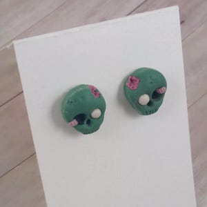 Könnte beinhalten: Ein Paar grüne Zombie-Schädel-Ohrringe mit rosa und weißen Details. Die Ohrringe sind aus Polymer Clay gefertigt und haben einen Durchmesser von etwa 2,5 cm.