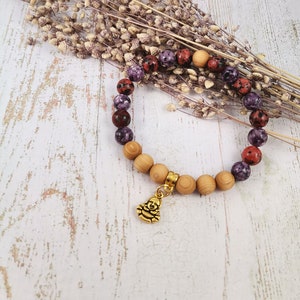 Könnte beinhalten: Ein Armband aus violetten, roten und braunen gesprenkelten Perlen und Holzperlen. Ein goldener Anhänger mit einem lächelnden Buddha ist am Armband befestigt.
