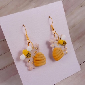 Könnte beinhalten: Ein Paar goldene Ohrhänger mit einem Design aus Honigwabe, Biene und Blattgold. Die Ohrringe sind aus Polymer Clay gefertigt und haben eine hellgelbe, weiße und goldene Farbe.