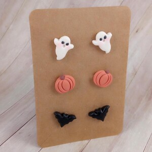 May include: A set of six Halloween-themed earrings. The earrings are made of polymer clay and feature a ghost, a pumpkin, and a bat. The earrings are displayed on a brown card.
