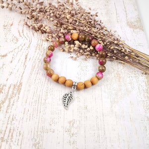 Könnte beinhalten: Ein Armband aus rosafarbenen und braunen Edelsteinen mit einem silbernen Blattanhänger. Das Armband besteht aus Holzperlen und liegt auf einem weißen Holzgrund.