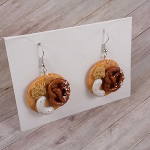Könnte beinhalten: Ein Paar Hängeohrringe mit einem Brezel, Senf und einer weißen Dipsauce auf einem Holzbrett. Die Ohrringe sind aus Polymer Clay gefertigt und haben silberne Haken.