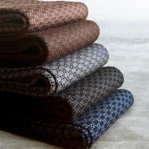 May include: A stack of five folded knit socks featuring a textured, repeating geometric pattern. The colours include shades of brown, grey, dark charcoal, and a deep blue, each with a distinct knit weave visible across the fabric surface.