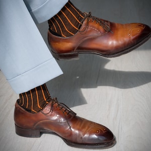 May include: A pair of brown leather dress shoes with intricate detailing on the toe, paired with brown socks featuring orange vertical stripes. The shoes are laced up and appear to be well-polished. The light blue pants are cuffed at the ankle.