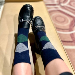 May include: A pair of black loafers with silver buckles, worn with dark blue socks featuring a green and grey geometric pattern.