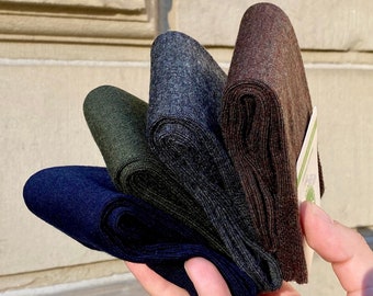 May include: A hand holds a fan of folded socks in shades of navy, olive green, charcoal, and brown. The socks are made of a textured material and appear to be new. A small tag is visible on the brown sock.