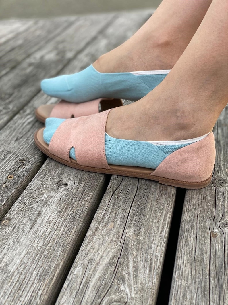 May include: A pair of light blue no-show socks with a white trim are worn with a pair of pink suede sandals. The sandals have a cut-out design and a flat sole.