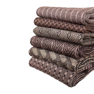 Puede incluir: Una pila de textiles de tela doblados en varios tonos marrones y neutros. Los textiles muestran una variedad de patrones, incluyendo rayas, cuadros y diseños geométricos. Las telas están cuidadosamente apiladas, mostrando las diferentes texturas y colores.