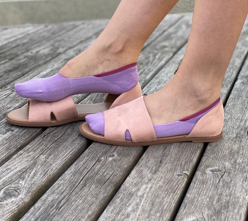 May include: A pair of lavender no-show socks with a pink trim are worn with a pair of open-toe sandals. The sandals are a light pink color and have a cut-out design.