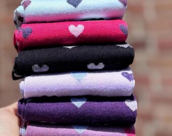 May include: A stack of folded socks in various colors, including light blue, pink, black, lavender, purple, and mauve. Each sock features a heart pattern. The socks are made of a soft, likely knit, material.