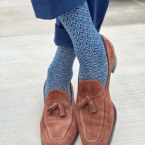 Puede incluir: Un par de mocasines de ante marrón con borlas y costuras visibles en la puntera, usados con calcetines de patrón geométrico en zigzag azul y azul claro, combinados con pantalones azul marino con dobladillo.