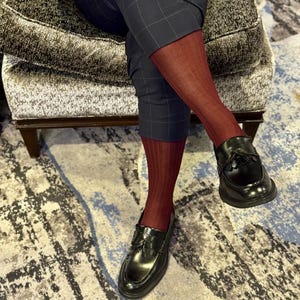 Op de afbeelding: Een persoon draagt ​​donker bordeauxrode geribbelde sokken en zwarte leren loafers met kwastjes. De persoon draagt ​​een marineblauwe geruite broek. De schoenen zijn glanzend en de sokken zijn effen.