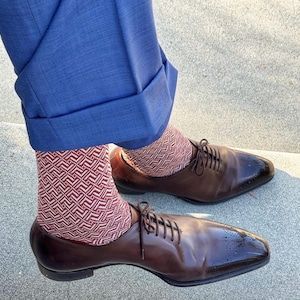 Puede incluir: Un par de zapatos oxford de cuero marrón oscuro con detalles brogue en la puntera, usados con calcetines de patrón geométrico rojo y blanco y pantalones azules con dobladillo. Los zapatos tienen cordones finos y un acabado pulido.