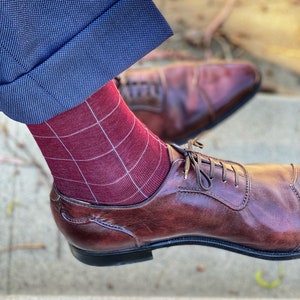 Op de afbeelding: Close-up van benen met bordeauxrode sokken met een wit ruitpatroon, gecombineerd met bruine leren schoenen. De blauwe pantalon is omgeslagen, wat een klassieke stijl geeft.