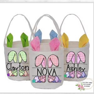 May include: Three white canvas Easter baskets with bunny ears. The baskets are decorated with pink, green, and blue gingham patterns. The baskets are personalized with the names "Clayton", "NOVA", and "Ashley".