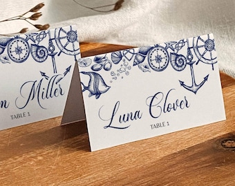 Può includere: Due segnaposto bianchi a tema nautico, con illustrazioni blu di conchiglie, ancore e bussole. Le carte mostrano i nomi "Adam Miller" e "Luna Clover" con "Table 1" sotto. Sono su un vassoio di legno.