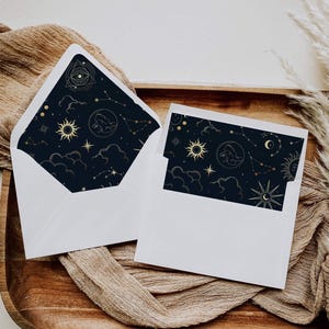 May include: Two white envelopes with dark navy blue liners featuring a gold celestial pattern. The design includes suns, moons, stars, constellations, clouds, and an eye within a triangle. The envelopes are open, displaying the patterned interior.