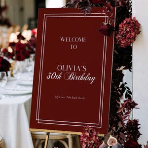 May include: A burgundy welcome sign on a gold easel, framed with white lines. The sign reads "WELCOME TO OLIVIA'S 50th Birthday" with the date and location below. The sign is surrounded by burgundy flowers and foliage.
