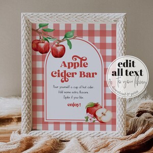 May include: A framed sign with the text "Apple Cider Bar" in a decorative font. The sign has a red and white gingham background and features watercolor illustrations of apples and flowers. The frame is white with a distressed finish.