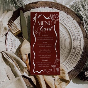 May include: A burgundy menu card with white script and illustrations, including a cupid and hearts. The menu lists starters, main courses, and desserts. The card is placed on a decorative plate with a fork and knife.