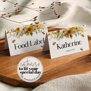 Puede incluir: Dos tarjetas de mesa blancas con detalles florales y de abeja. Una dice "Food Label" y la otra "Katherine, Table One". Un gráfico circular dice "Personaliza para tu día especial, edita, descarga e imprime."