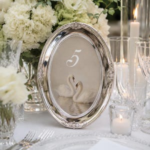 May include: An oval silver table number holder with a decorative border. The number "5" is written in white script above a painting of two swans. The table setting includes white flowers, candles, and glassware.