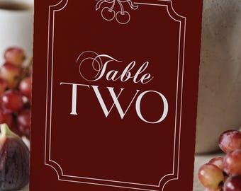 May include: A burgundy table card with white text that reads "Table TWO." The card has a decorative white border and a small bow and cherry design at the top. Grapes and figs are visible in the background.