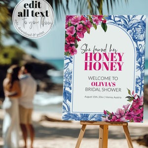 May include: A white sign with blue and pink floral borders. The sign reads "She found her HONEY HONEY WELCOME TO OLIVIA'S BRIDAL SHOWER August 15th, 20xx | Vienna, Austria".