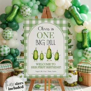 Op de afbeelding: Een houten ezel toont een bord met een groen-witte geruite rand. Op het bord staat "Olivia is ONE BIG DILL" met cartoon augurken illustraties. Groene ballonnen en augurk-vormige ballonnen versieren de achtergrond. Potten met augurken zijn ook zichtbaar.