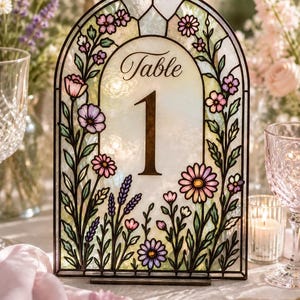 Puede incluir: Número de mesa de vidriera con diseño floral. El letrero arqueado presenta un fondo color crema con la palabra "Table" y el número "1" en marrón. Flores rosas, moradas y amarillas y hojas verdes decoran los bordes.