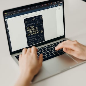 May include: A laptop computer displaying a wedding invitation with a dark blue background and gold stars, moons, and clouds. The invitation text reads "Please join us as we celebrate the marriage of Olivia Harrison and Michael Hendricks".