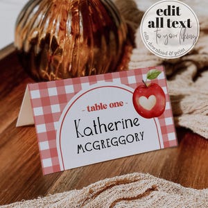 May include: A folded place card with a red and white gingham pattern. The card features the text "table one", the name "Katherine MCGREGGORY", and a watercolor apple illustration with a heart-shaped core. The card also has the text "edit all text to your liking".