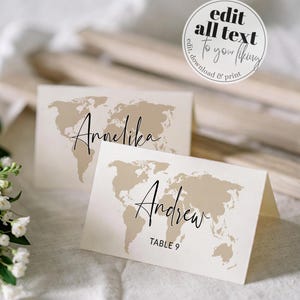 Può includere: Due segnaposto piegati con un design a mappa del mondo beige. Le carte hanno nomi scritti in corsivo nero: "Annelika" e "Andrew". La seconda carta ha anche "TABLE 9" stampato sotto il nome. Una grafica circolare recita "EDIT all text To your liking edit, download & print".