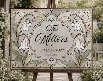 Può includere: Un cartello rettangolare in vetro colorato con un motivo floreale di fiori bianchi a forma di campana e foglie verdi. Il centro contiene il testo The Millers, OLIVIA & ADAM e 03.03.20XX. Il cartello è montato su un cavalletto in legno.