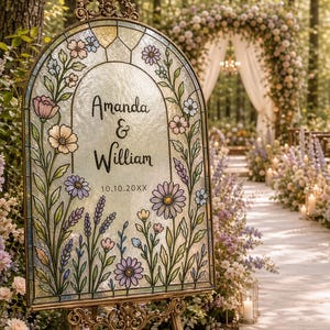 Pode incluir: Uma placa de casamento em vitral com os nomes "Amanda & William" e a data "10.10.20XX". A placa &eacute; decorada com flores e folhagens coloridas, colocada em um cavalete dourado, em um cen&aacute;rio de jardim.