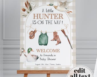 May include: A rectangular sign featuring a beige and white checkered border with a central arch. It displays a watercolour mallard duck, cattails, a hat, green overalls, boots, and a rifle hanging on a line. Text reads: A little HUNTER IS ON THE WAY! WELCOME to Amanda's Baby Shower, August 15th, 20XX | Vienna, Austria.
