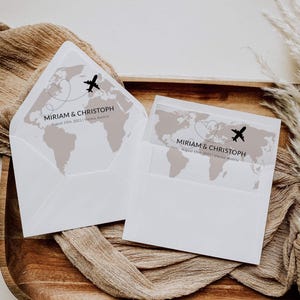 May include: Two white wedding invitation envelopes with a world map design and a heart-shaped dotted line with a small airplane. The text "MIRIAM & CHRISTOPH August 15th, 2021 | Vienna, Austria" is printed on the envelopes. The envelopes are on a wooden tray.